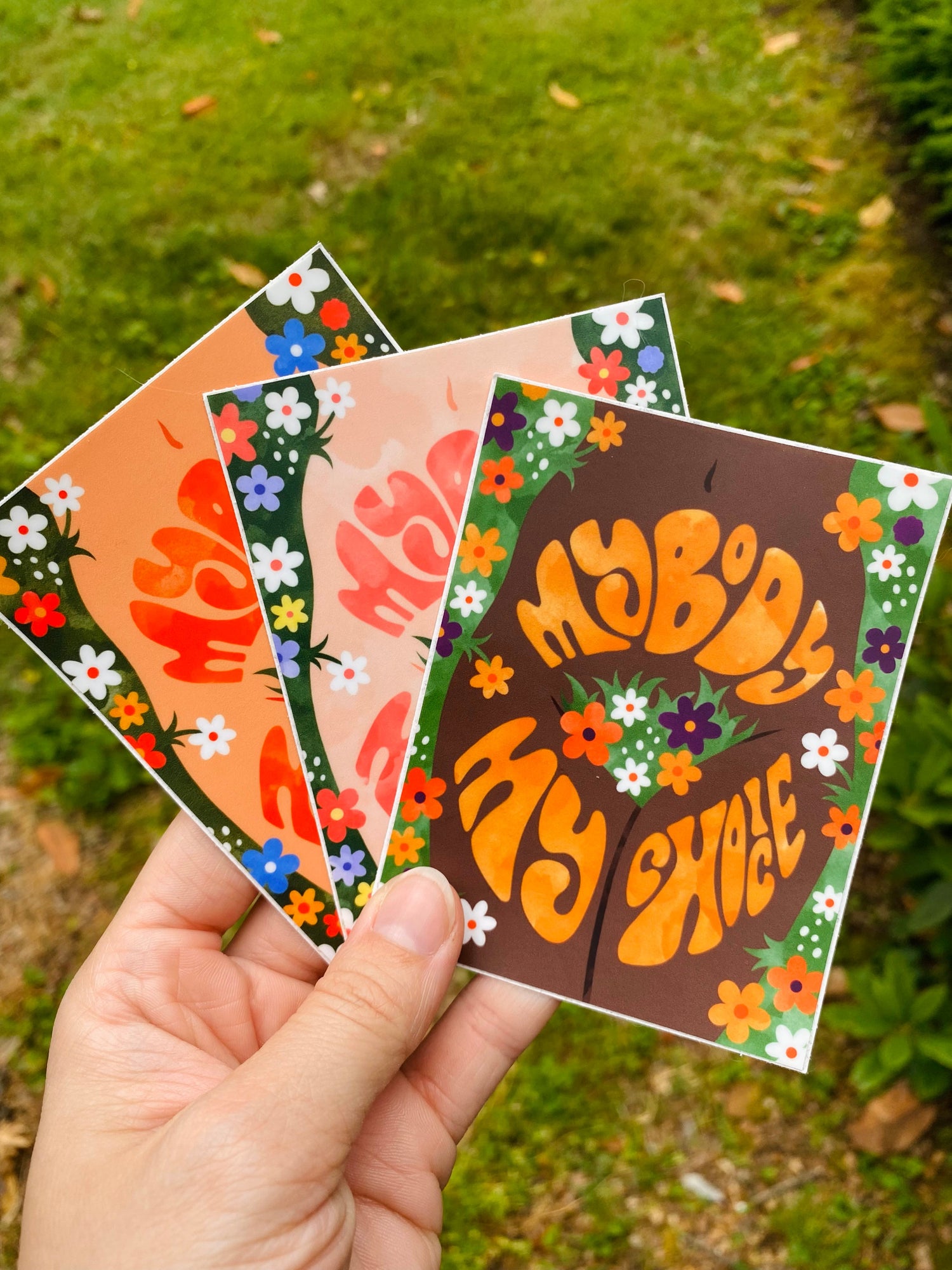 a hand holding three stickers, all variations of the same illustration showing a woman's body surrounded by flowers with the phrase 'my body my choice'